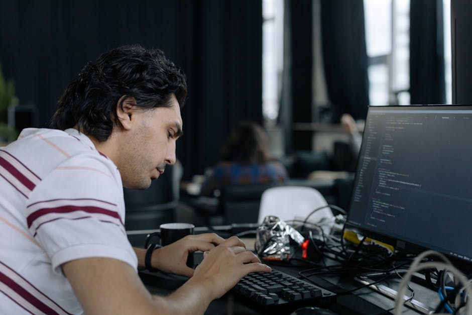 A young man with dark, wavy hair wearing a white polo shirt with red and gray stripes is seated at a desk in an indoor office or educational setting, focused on programming on his computer screen. The