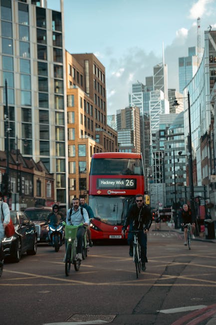 A busy urban street scene featuring modern high-rise buildings with glass facades and brick exteriors under a partly cloudy sky. In the foreground, several pedestrians are walking and cycling along th